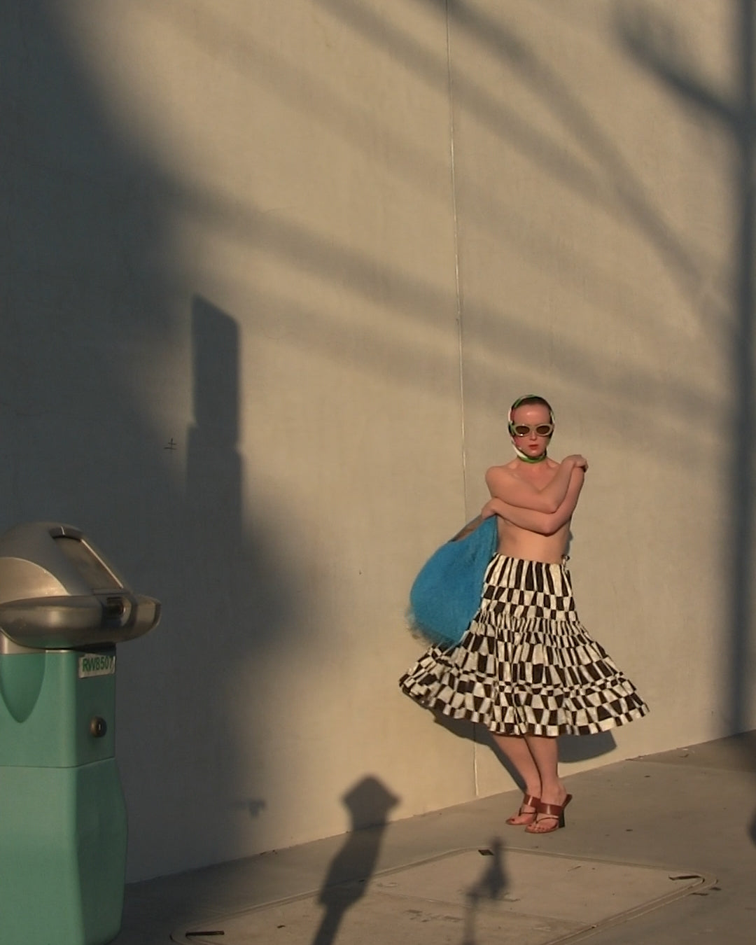 Woman in a checkered skirt and sunglasses standing against a wall with a shadow. 