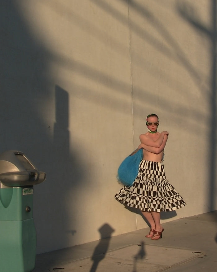 Woman in a checkered skirt and sunglasses standing against a wall with a shadow. 