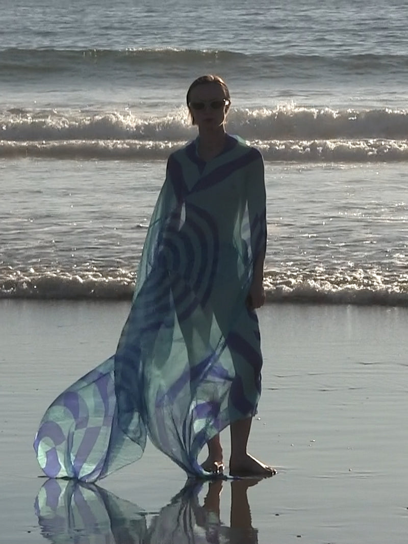 Person wearing a flowing blue patterned sheer dress standing barefoot at the shoreline, with ocean waves and reflections on the wet sand