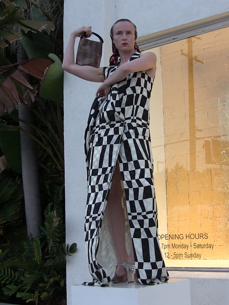 Person posing in a black-and-white geometric patterned dress while holding a small handbag, standing in front of a storefront window