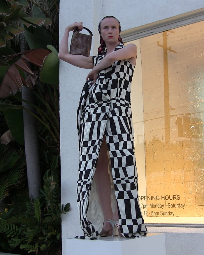 Person posing in a black-and-white geometric patterned dress while holding a small handbag, standing in front of a storefront window