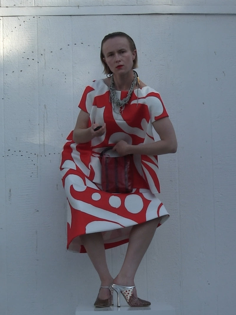 Woman in a bold red-and-white patterned dress posing against the white wall, accessorized with layered silver necklace, pointed heels and a small handbag 