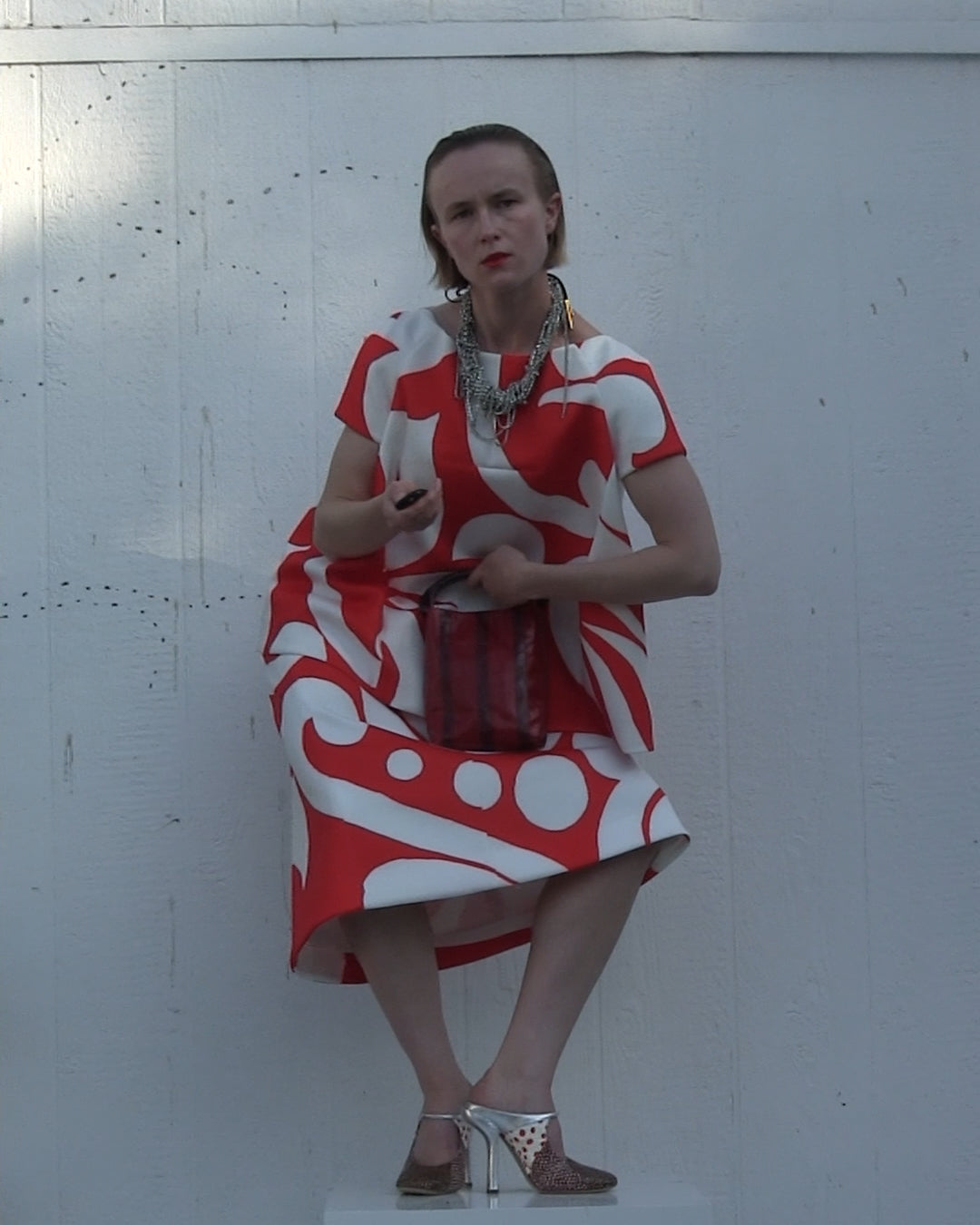 Woman in a bold red-and-white patterned dress posing against the white wall, accessorized with layered silver necklace, pointed heels and a small handbag 