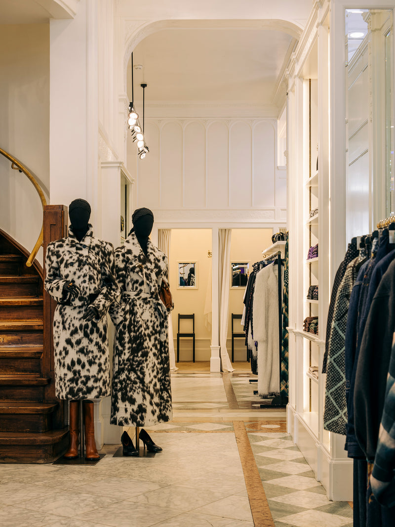 Two mannequins wearing matching black and white patterned long coats stand in a boutique interior with a staircase on the left and clothing racks on the right.
