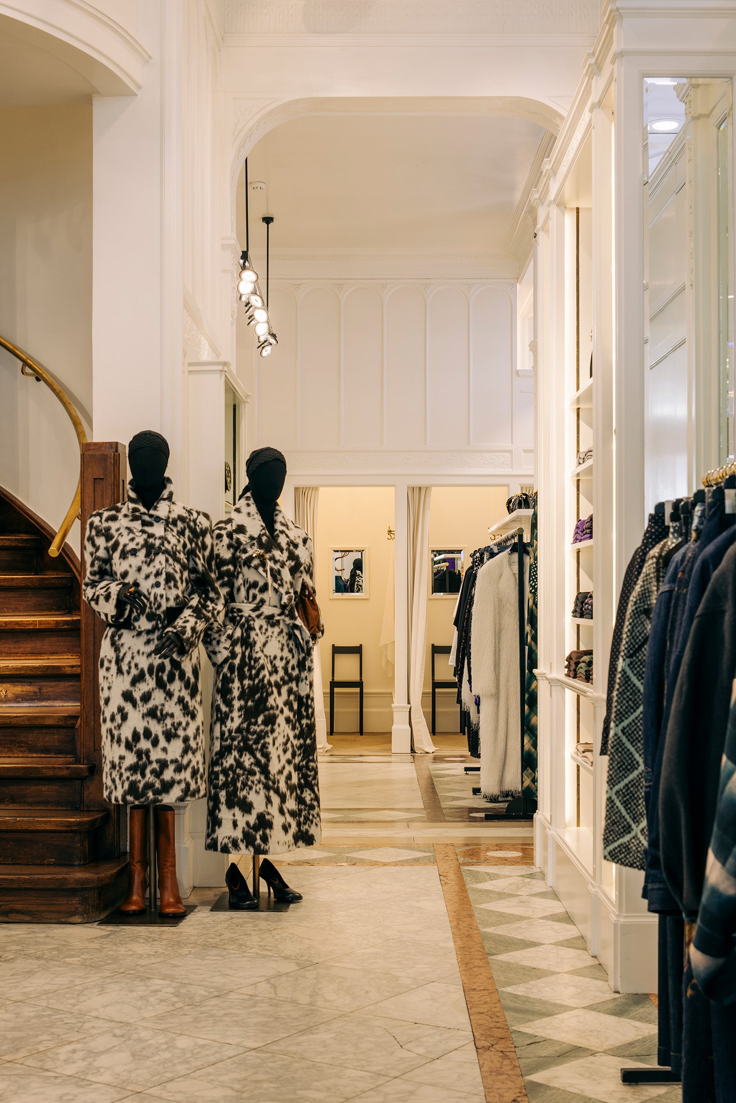 Two mannequins wearing matching black and white patterned long coats stand in a boutique interior with a staircase on the left and clothing racks on the right.