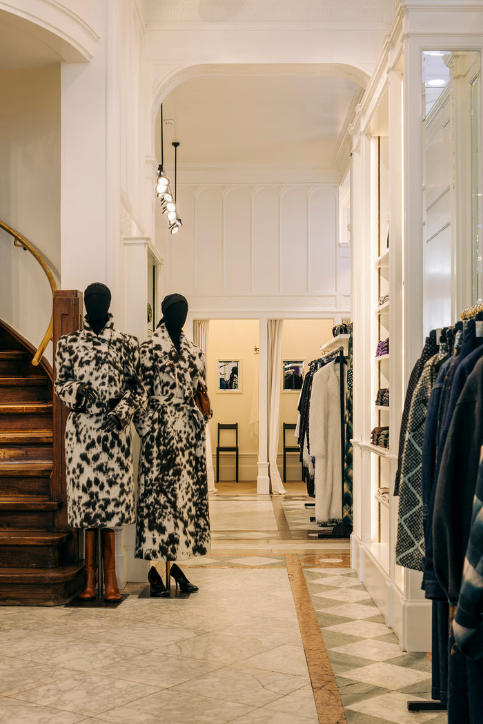 Two mannequins wearing matching black and white patterned long coats stand in a boutique interior with a staircase on the left and clothing racks on the right.