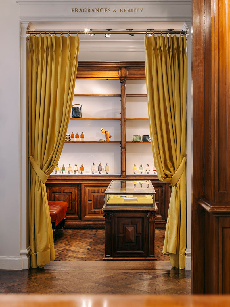 A luxurious interior view of a fragrance and beauty, framed by heavy mustard yellow curtains tied back on either side. Through the doorway, a dark wood display cabinet and illuminated shelves filled with bottles are visible.