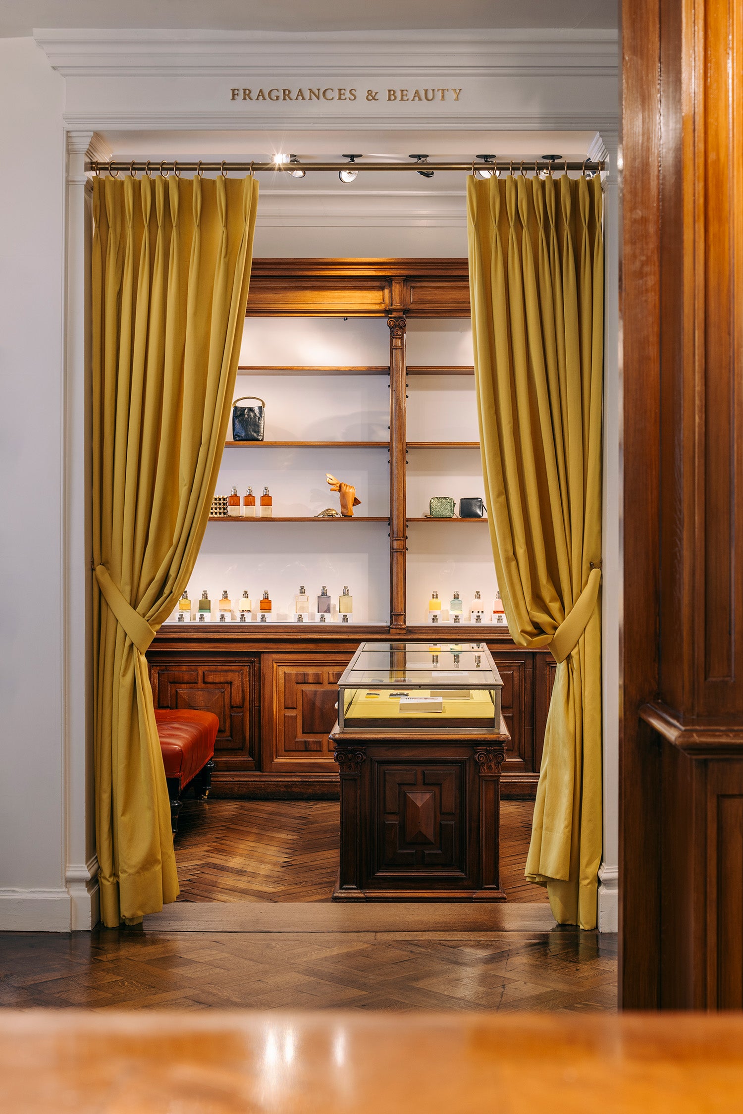 A luxurious interior view of a fragrance and beauty, framed by heavy mustard yellow curtains tied back on either side. Through the doorway, a dark wood display cabinet and illuminated shelves filled with bottles are visible.
