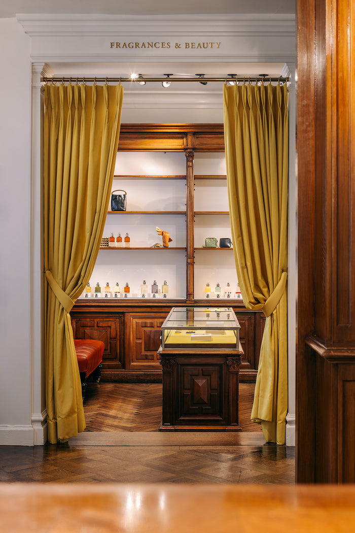 A luxurious interior view of a fragrance and beauty, framed by heavy mustard yellow curtains tied back on either side. Through the doorway, a dark wood display cabinet and illuminated shelves filled with bottles are visible.