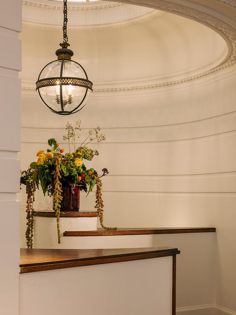 A vintage-style hanging globe chandelier with a black metal frame and clear glass globe, suspended in a cream-colored domed interior with wooden railings and a floral arrangement.