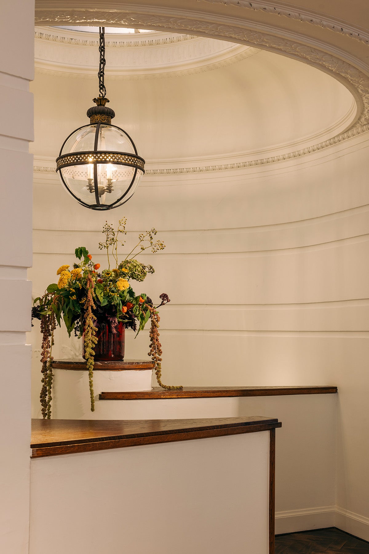 A vintage-style hanging globe chandelier with a black metal frame and clear glass globe, suspended in a cream-colored domed interior with wooden railings and a floral arrangement.