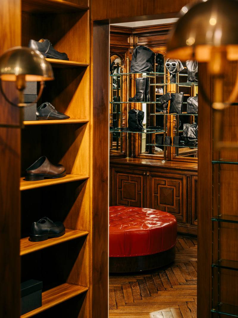 A warm, upscale interior scene showing a wooden shelving unit on the left with several pairs of shoes, a red tufted ottoman in the center, and a mirrored closet area with black garments in the background. 