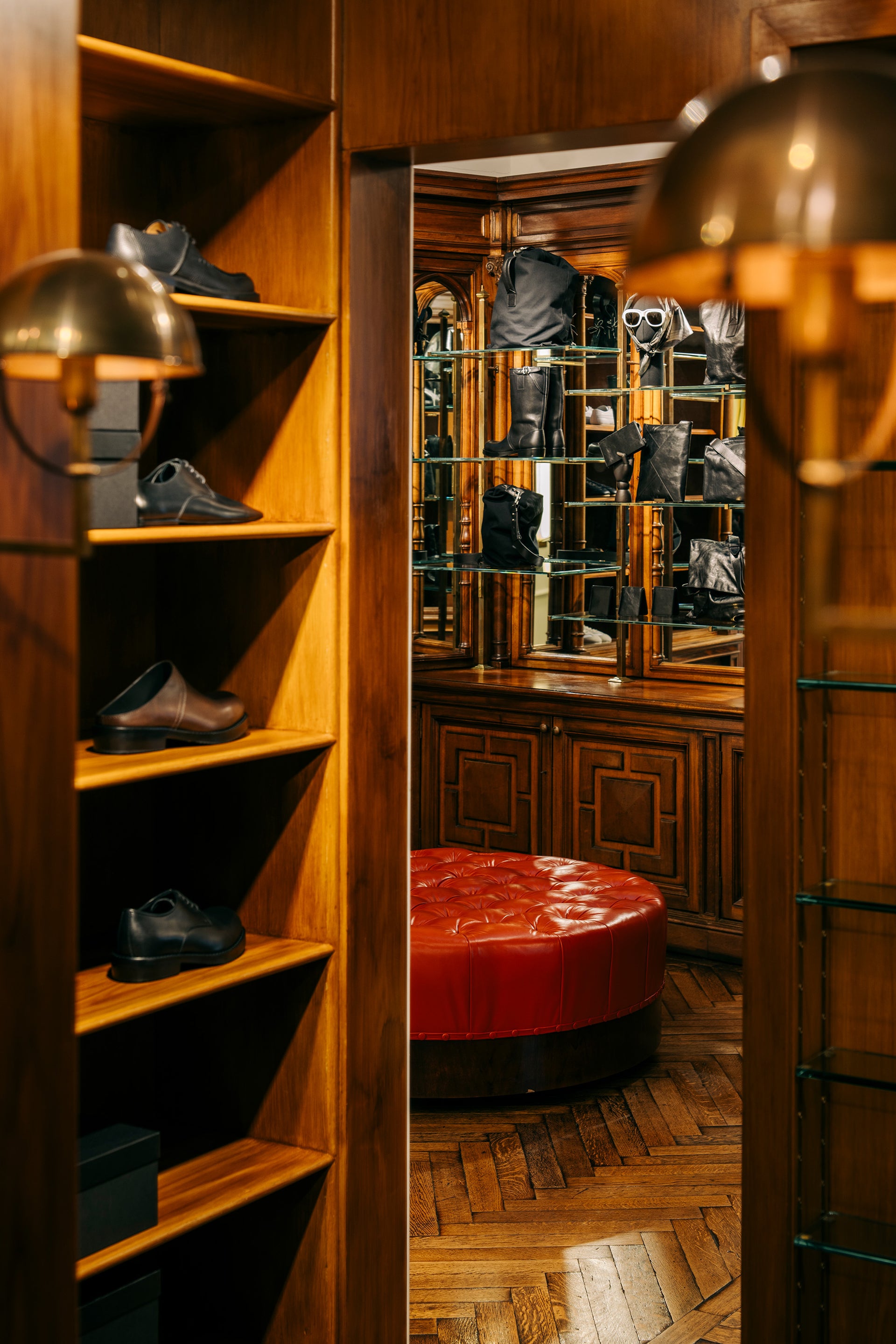A warm, upscale interior scene showing a wooden shelving unit on the left with several pairs of shoes, a red tufted ottoman in the center, and a mirrored closet area with black garments in the background. 