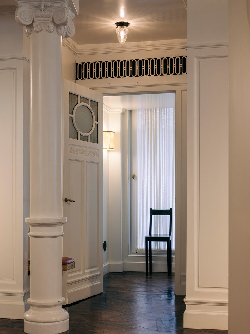 An interior hallway with white paneled walls and columns, a dark wood floor, a black chair near a window with white curtains, and a decorative black-and-white grille above the doorway.