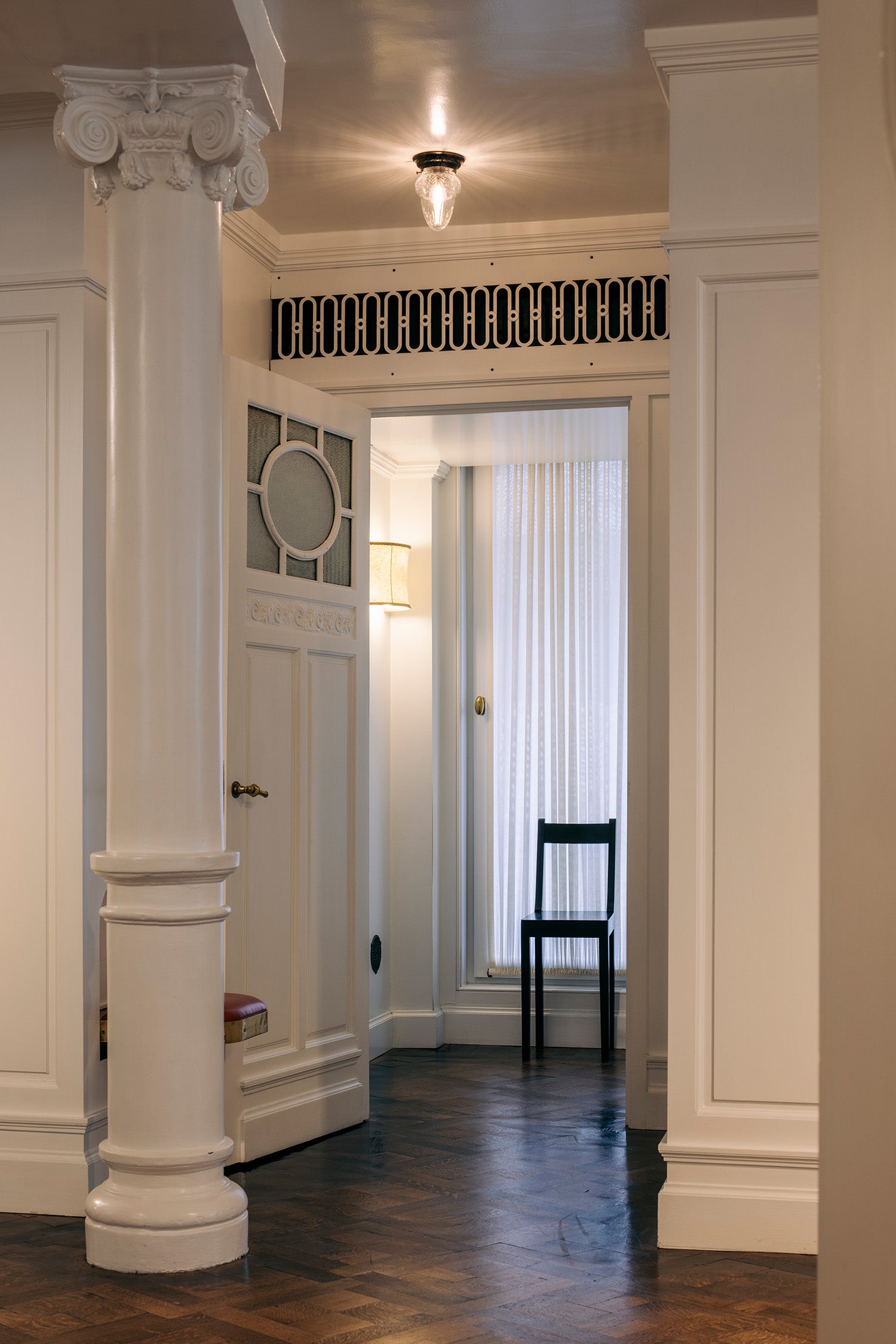 An interior hallway with white paneled walls and columns, a dark wood floor, a black chair near a window with white curtains, and a decorative black-and-white grille above the doorway.