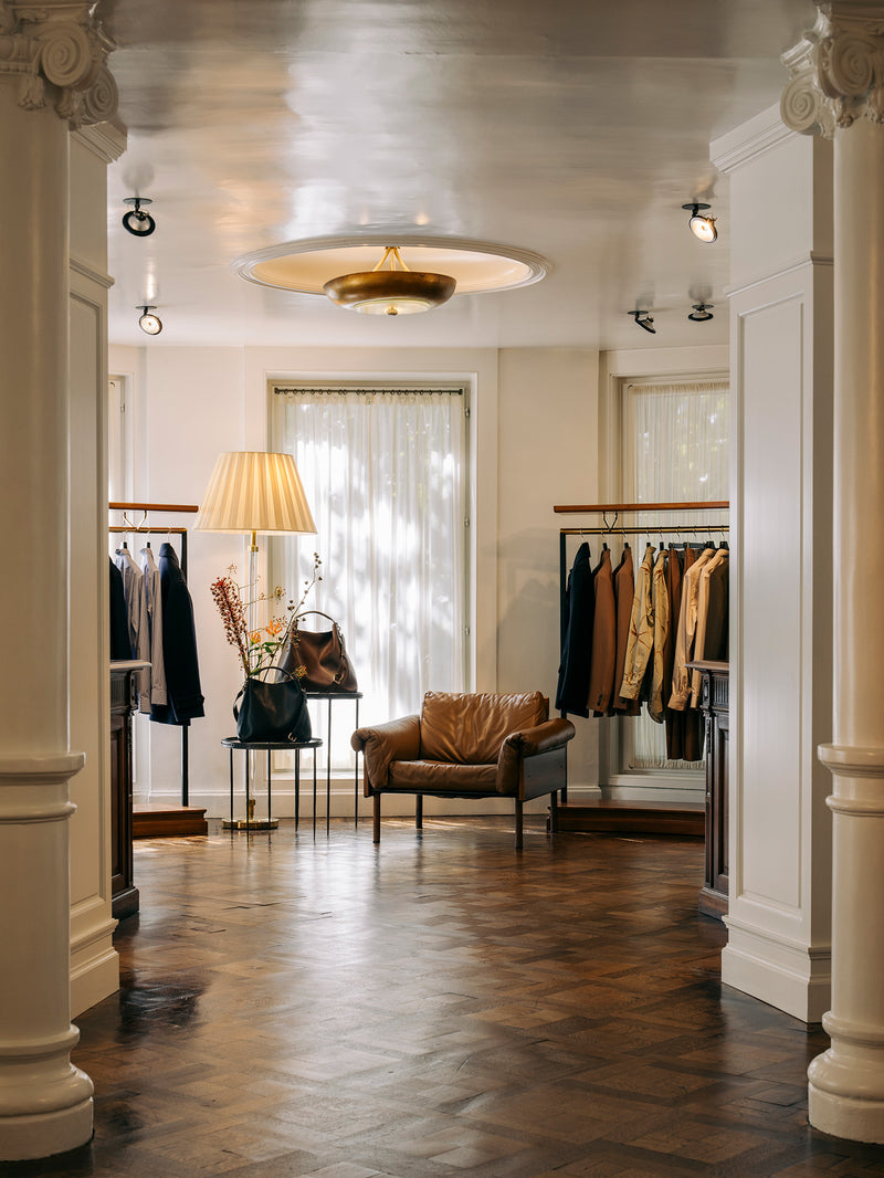 A boutique interior with a brown leather armchair centered in a hallway, clothing racks with garments, a floor lamp, and a warm wood parquet floor. 