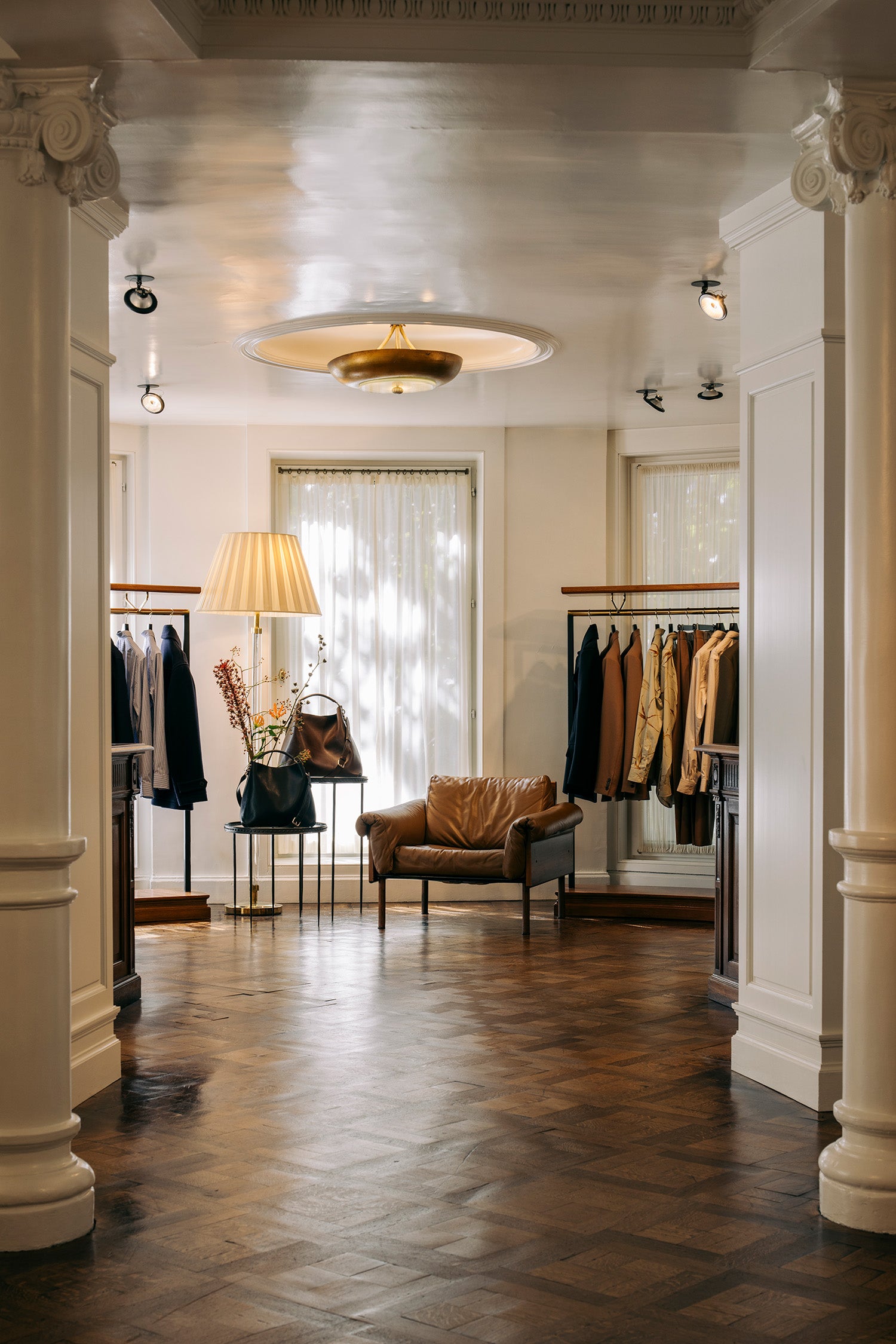 A boutique interior with a brown leather armchair centered in a hallway, clothing racks with garments, a floor lamp, and a warm wood parquet floor. 