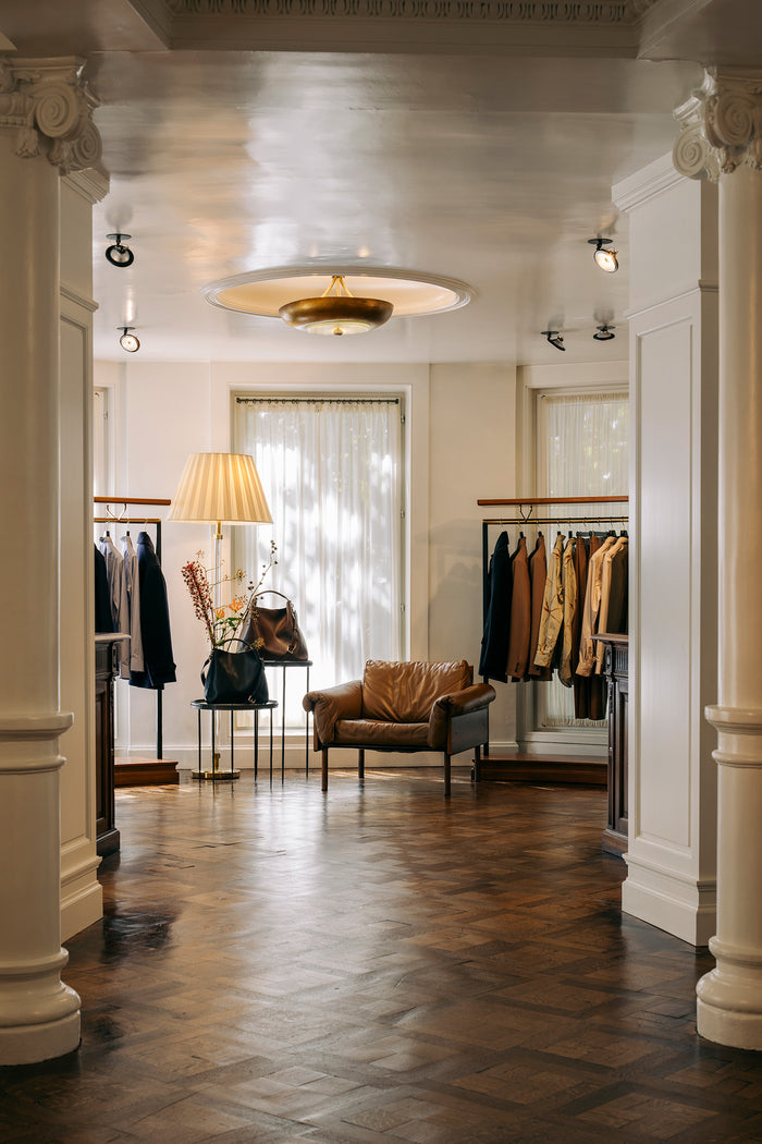 A boutique interior with a brown leather armchair centered in a hallway, clothing racks with garments, a floor lamp, and a warm wood parquet floor. 