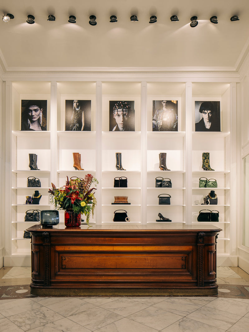 A luxury boutique interior featuring a large dark wood counter in front of white built-in shelving. The shelves display various black leather handbags, boots, and shoes, with framed black-and-white fashion portraits mounted above.