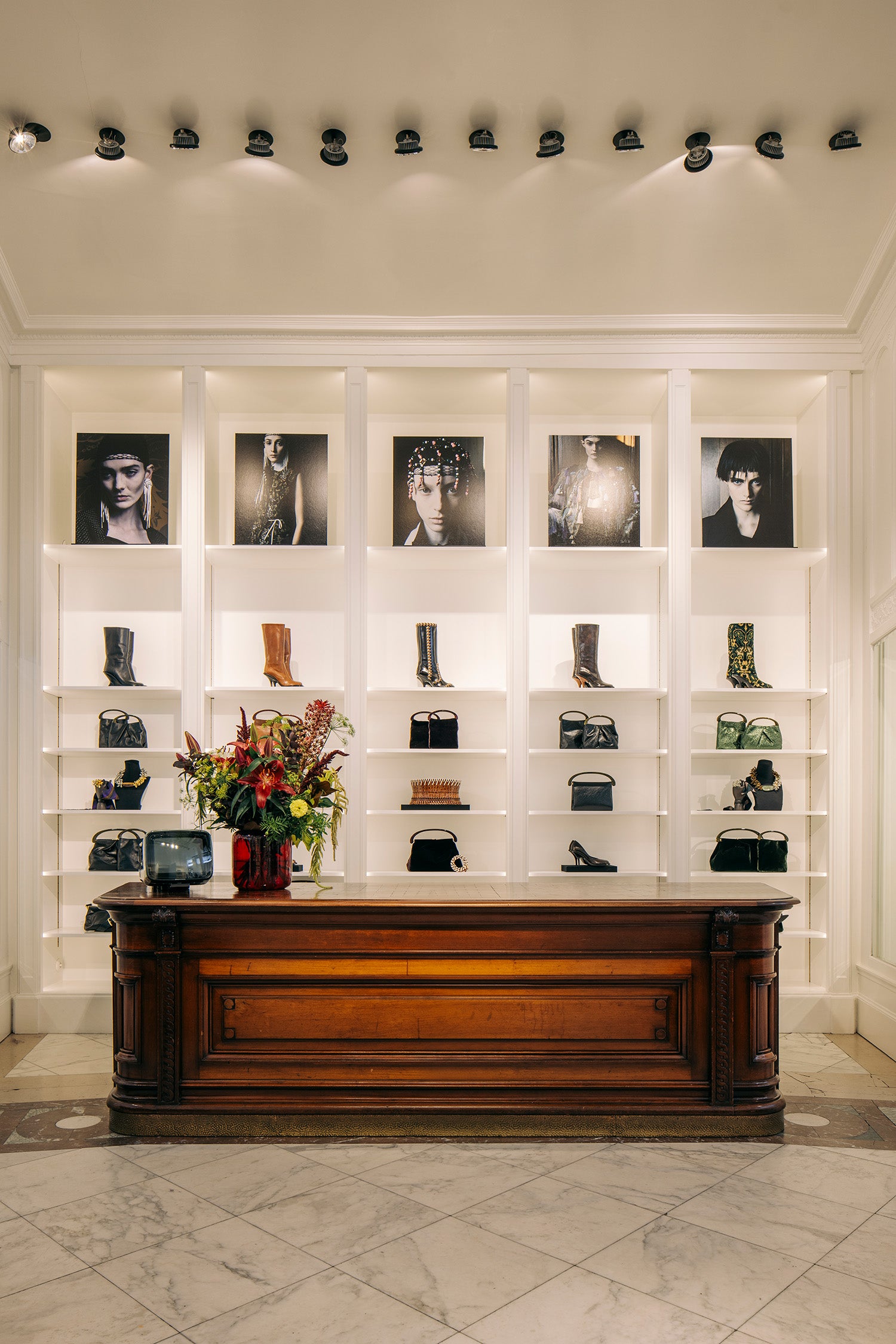 A luxury boutique interior featuring a large dark wood counter in front of white built-in shelving. The shelves display various black leather handbags, boots, and shoes, with framed black-and-white fashion portraits mounted above.