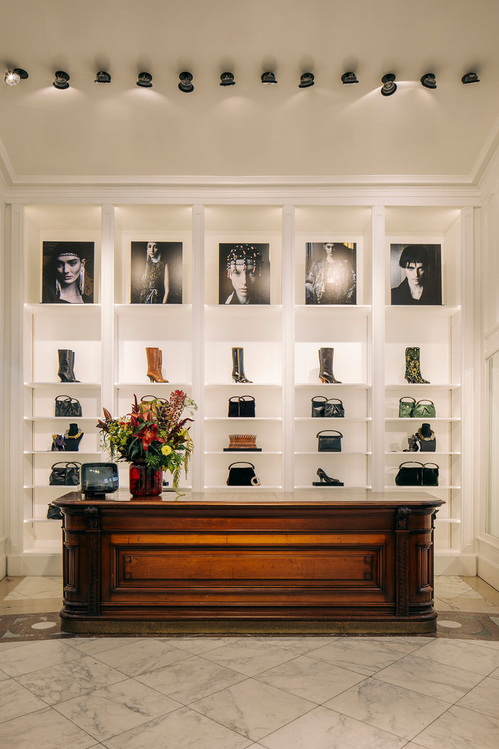 A luxury boutique interior featuring a large dark wood counter in front of white built-in shelving. The shelves display various black leather handbags, boots, and shoes, with framed black-and-white fashion portraits mounted above.