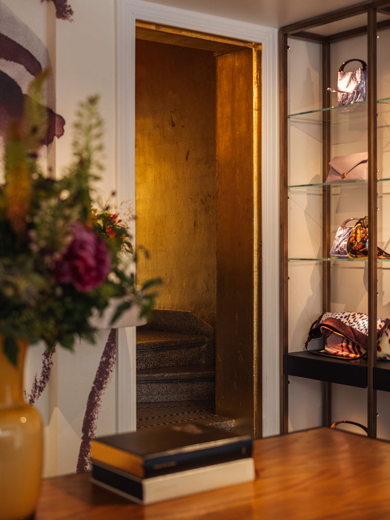 A warm, stylish interior scene featuring a gold-framed doorway, a wooden table with books and a vase of flowers in the foreground, and a tall glass display cabinet on the right holding fashion accessories.