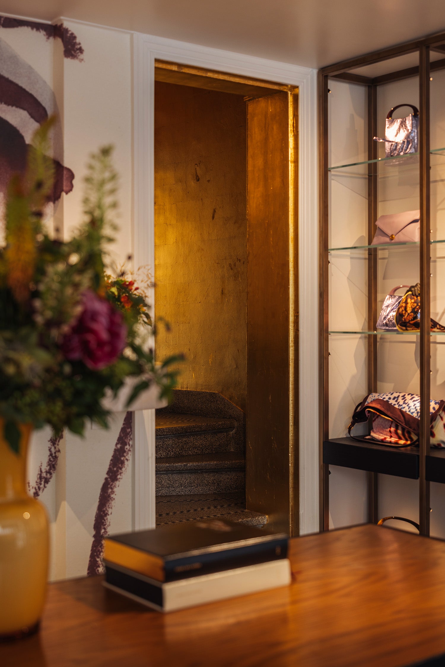 A warm, stylish interior scene featuring a gold-framed doorway, a wooden table with books and a vase of flowers in the foreground, and a tall glass display cabinet on the right holding fashion accessories.