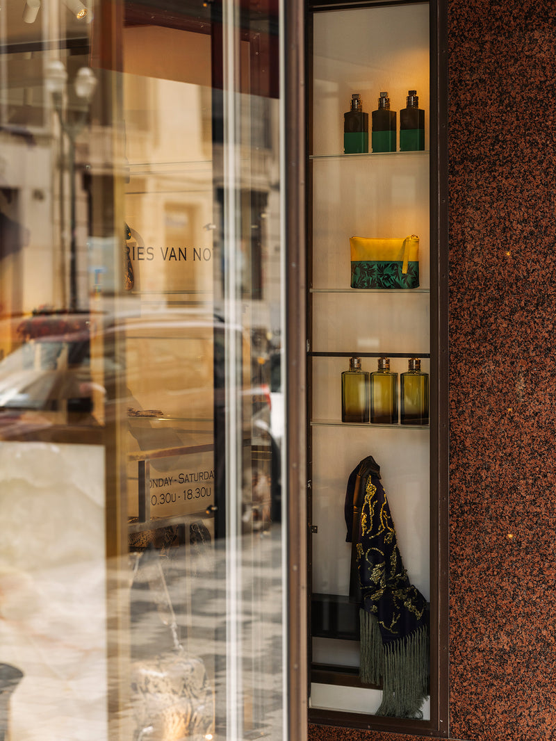 A storefront display cabinet with glass doors and shelves, showcasing various bottles and containers. The cabinet is set against a textured brown speckled wall, with reflections of the street and shop interior visible on the left glass pane.