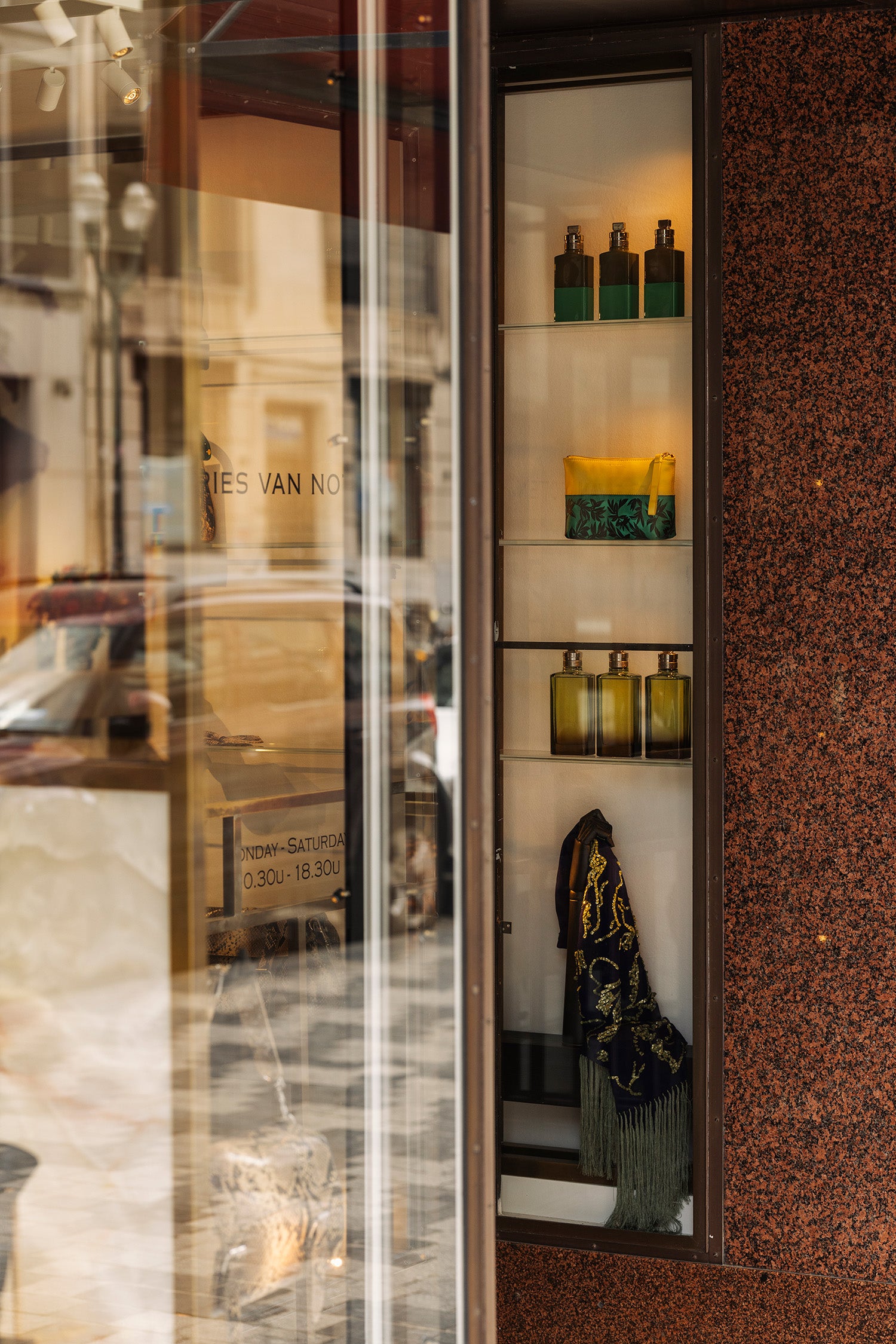 A storefront display cabinet with glass doors and shelves, showcasing various bottles and containers. The cabinet is set against a textured brown speckled wall, with reflections of the street and shop interior visible on the left glass pane.
