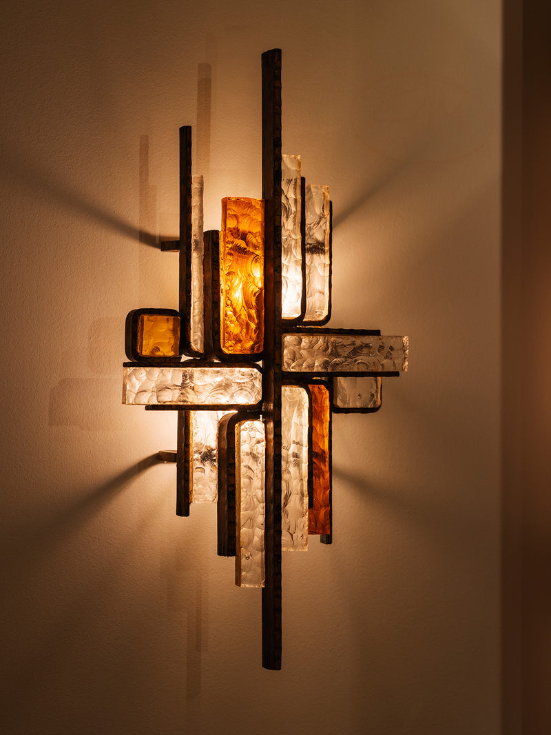 An abstract wall sconce light fixture with intersecting vertical and horizontal bars, featuring textured clear glass panels and amber-colored glass inserts, casting a warm glow on a beige wall.