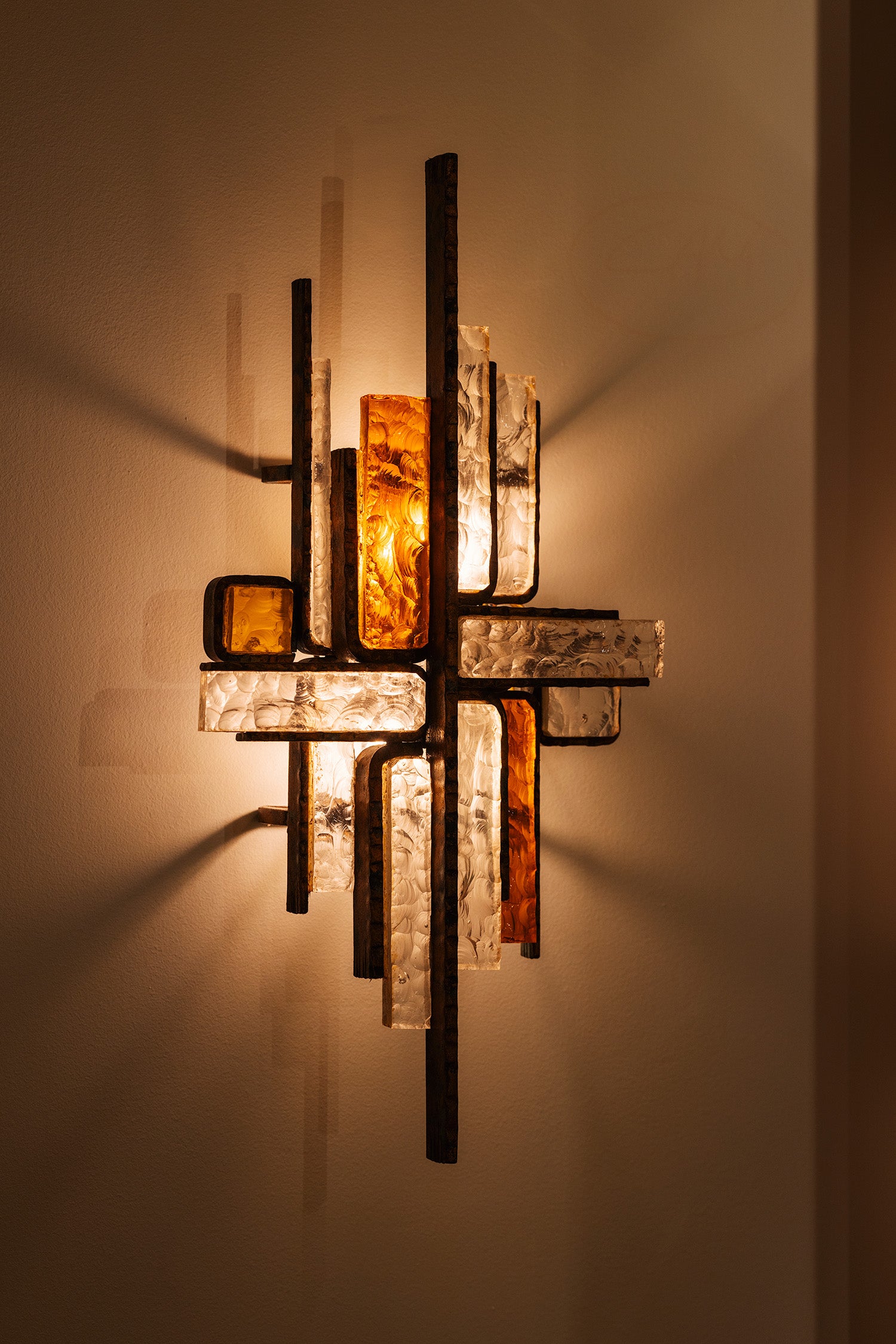 An abstract wall sconce light fixture with intersecting vertical and horizontal bars, featuring textured clear glass panels and amber-colored glass inserts, casting a warm glow on a beige wall.