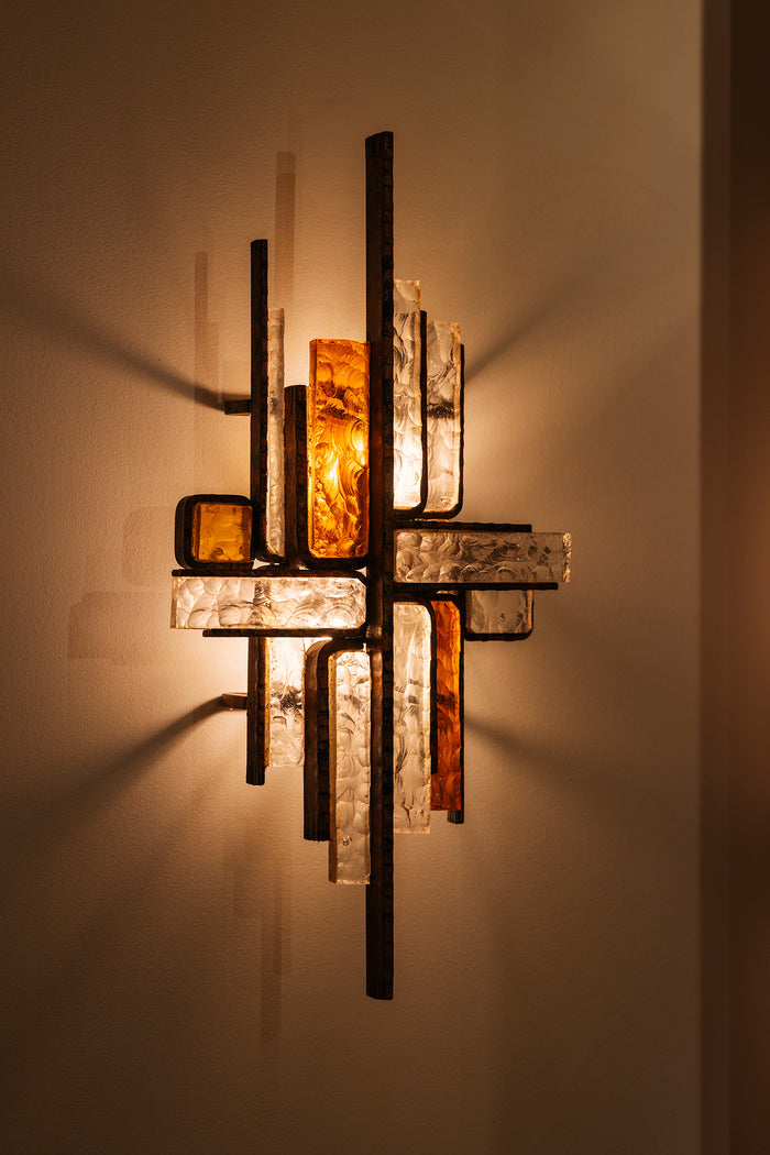 An abstract wall sconce light fixture with intersecting vertical and horizontal bars, featuring textured clear glass panels and amber-colored glass inserts, casting a warm glow on a beige wall.
