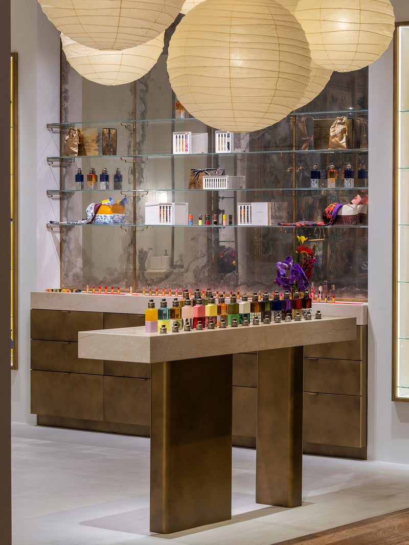 A display of numerous small colorful bottles arranged on a marble-topped counter in a luxury interior, with large cream paper lanterns hanging overhead and mirrored shelving in the background.