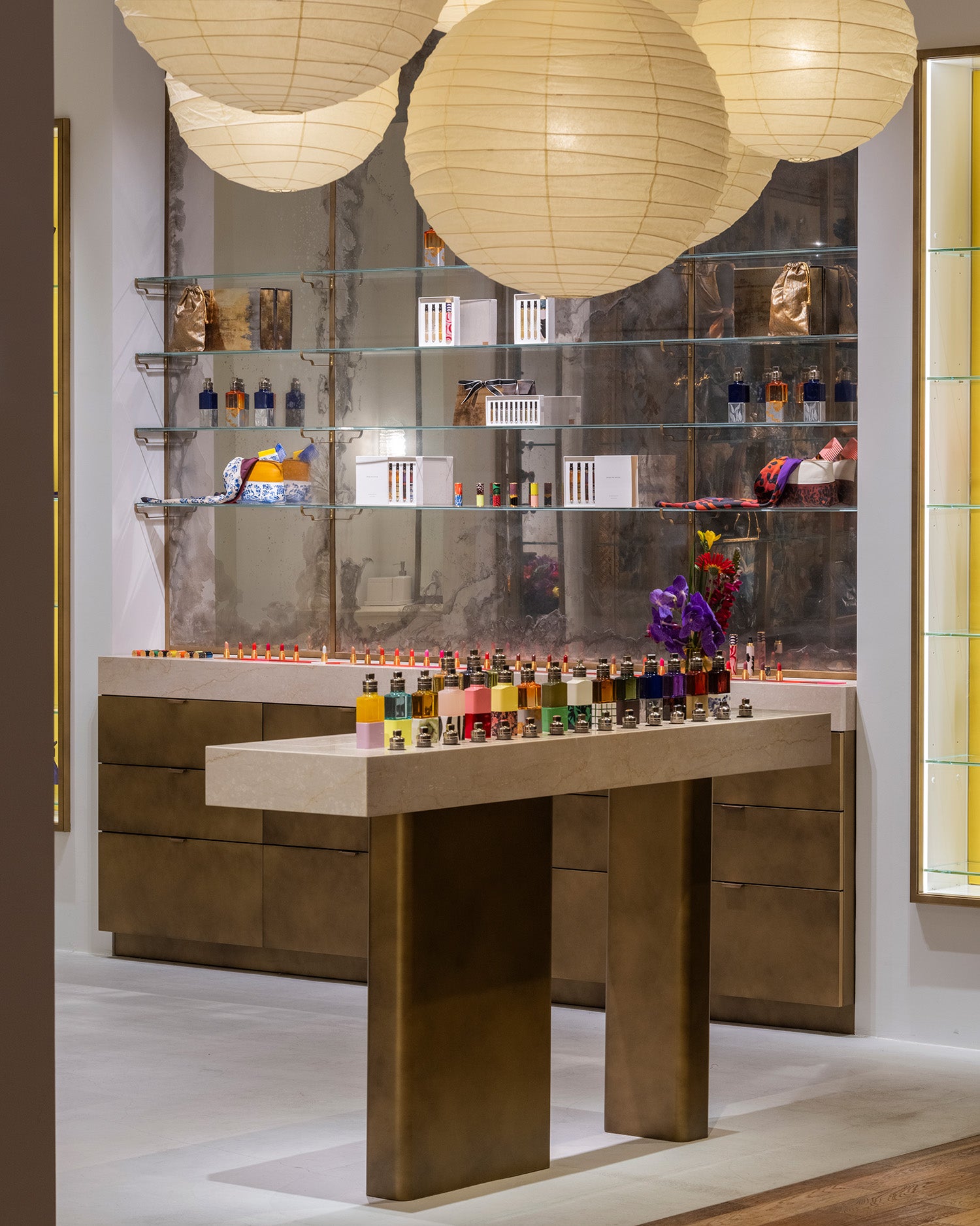 A display of numerous small colorful bottles arranged on a marble-topped counter in a luxury interior, with large cream paper lanterns hanging overhead and mirrored shelving in the background.
