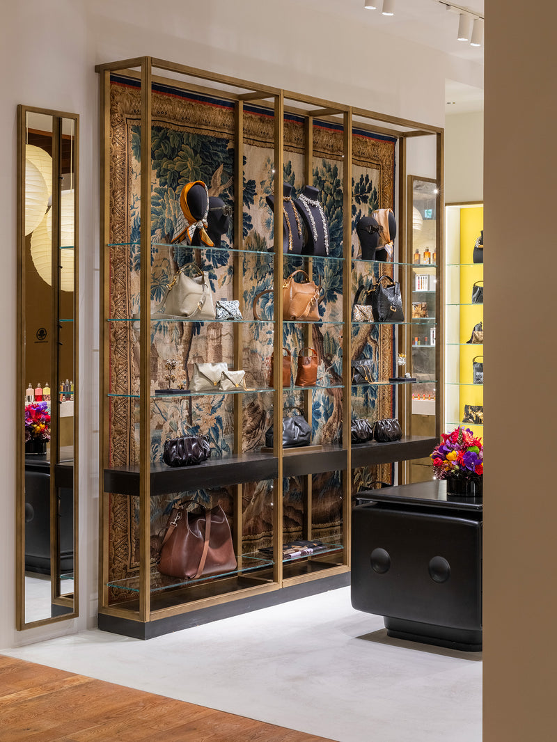 A luxury retail display cabinet featuring a large blue and gold patterned tapestry backdrop, gold-framed glass shelves, and mannequins displaying fashion items, set in a modern boutique interior.