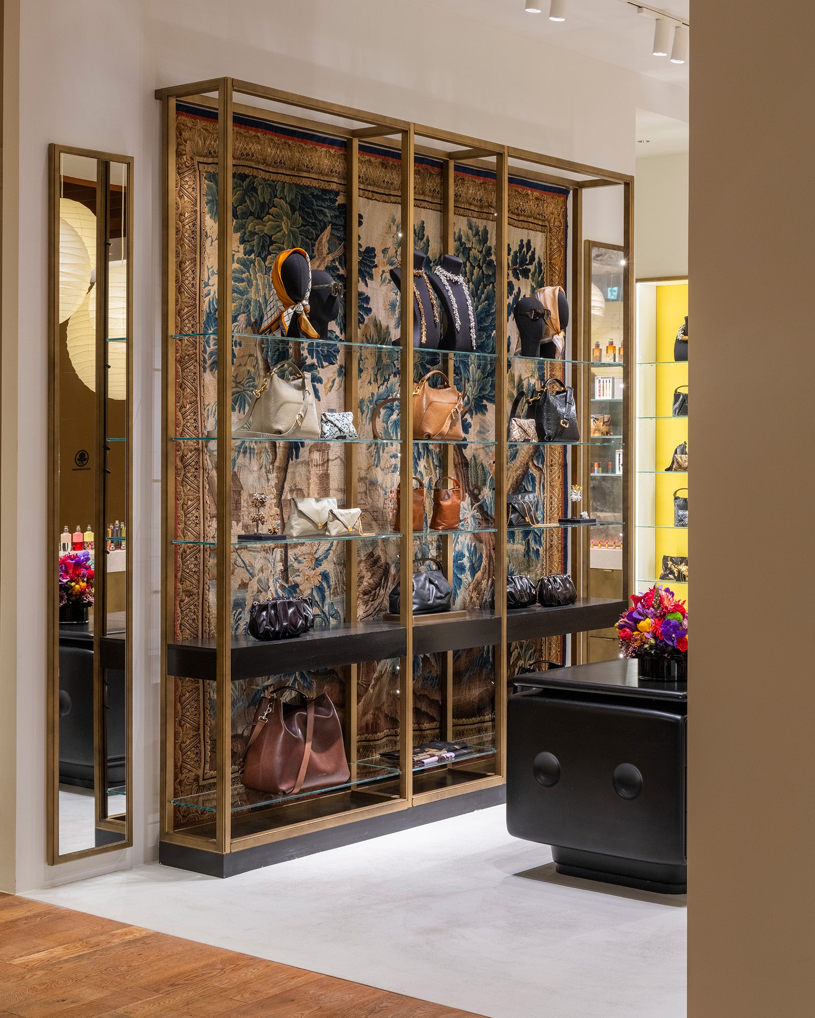 A luxury retail display cabinet featuring a large blue and gold patterned tapestry backdrop, gold-framed glass shelves, and mannequins displaying fashion items, set in a modern boutique interior.