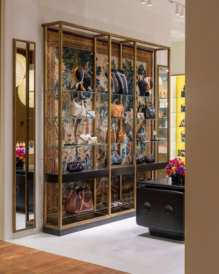 A luxury retail display cabinet featuring a large blue and gold patterned tapestry backdrop, gold-framed glass shelves, and mannequins displaying fashion items, set in a modern boutique interior.