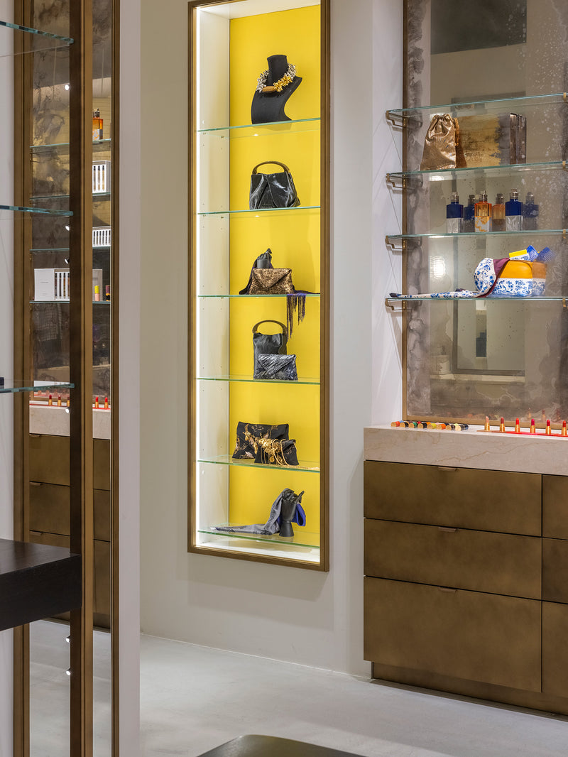 A vertical retail display case with a bright yellow backlit wall and six glass shelves, showcasing multiple handbags and accesories. 