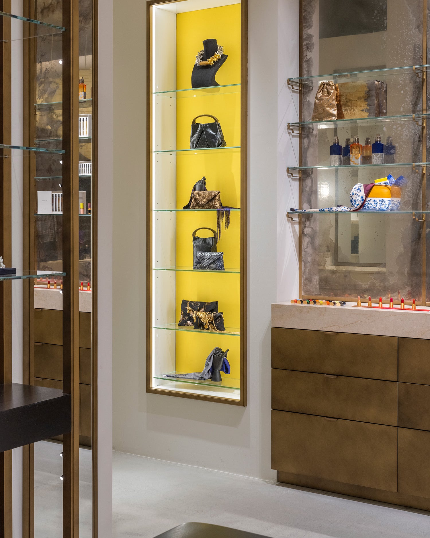 A vertical retail display case with a bright yellow backlit wall and six glass shelves, showcasing multiple handbags and accesories. 