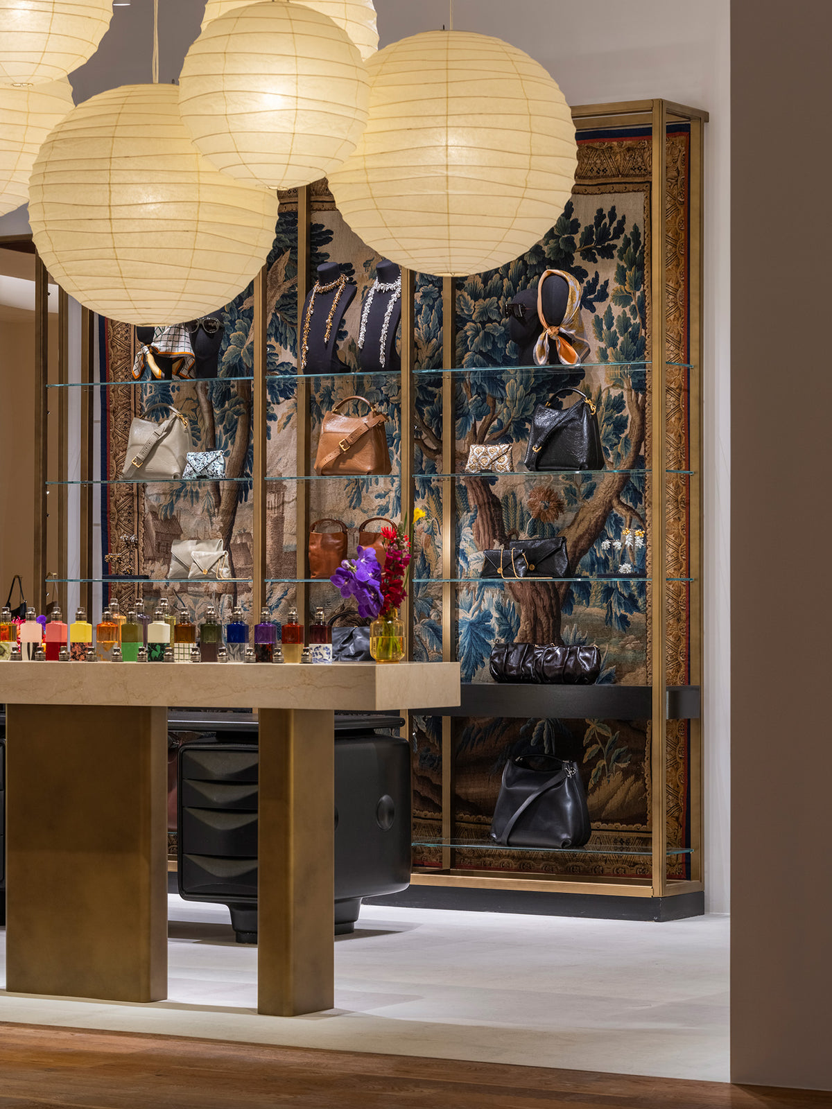 A luxury interior display featuring a large glass display cabinet with an ornate blue and gold tapestry background, showcasing handbags and accessories. In the foreground, a wooden table with gold-toned metal legs holds colorful fragrance bottles and a small yellow vase with purple flowers.