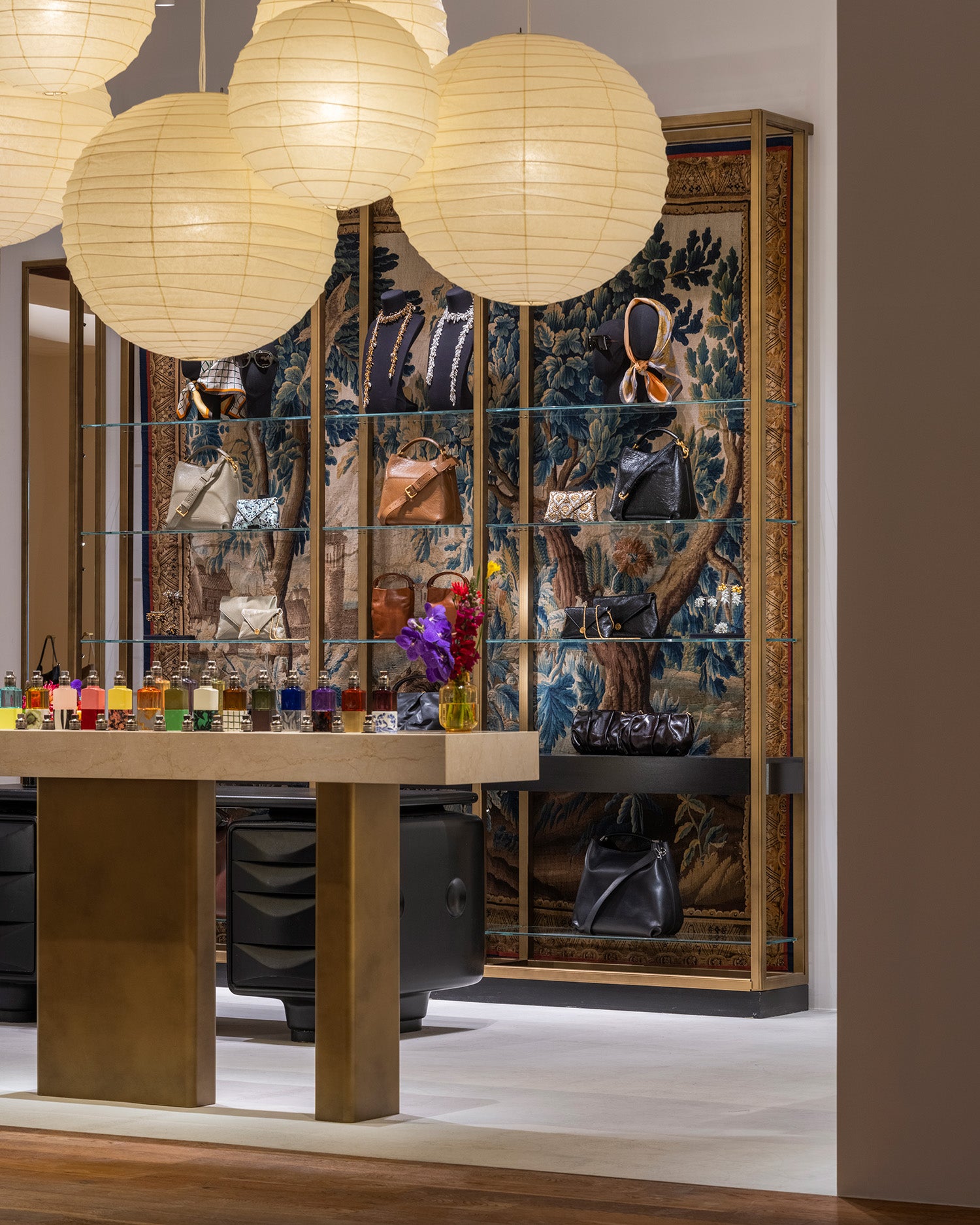 A luxury interior display featuring a large glass display cabinet with an ornate blue and gold tapestry background, showcasing handbags and accessories. In the foreground, a wooden table with gold-toned metal legs holds colorful fragrance bottles and a small yellow vase with purple flowers.