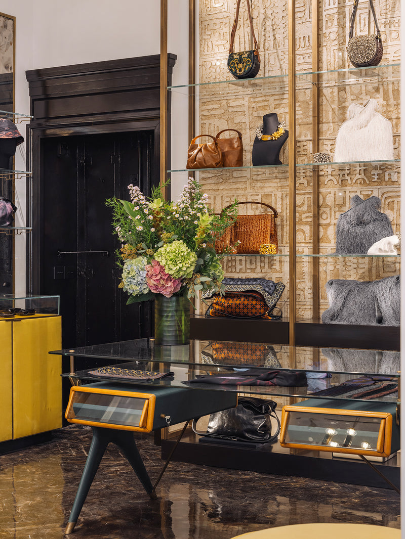 A luxury boutique interior featuring a glass display table with orange-trimmed drawers, a bouquet, and a wall of gold-patterned shelving displaying handbags, accessories and jewelry.