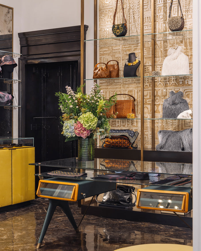A luxury boutique interior featuring a glass display table with orange-trimmed drawers, a bouquet, and a wall of gold-patterned shelving displaying handbags, accessories and jewelry.