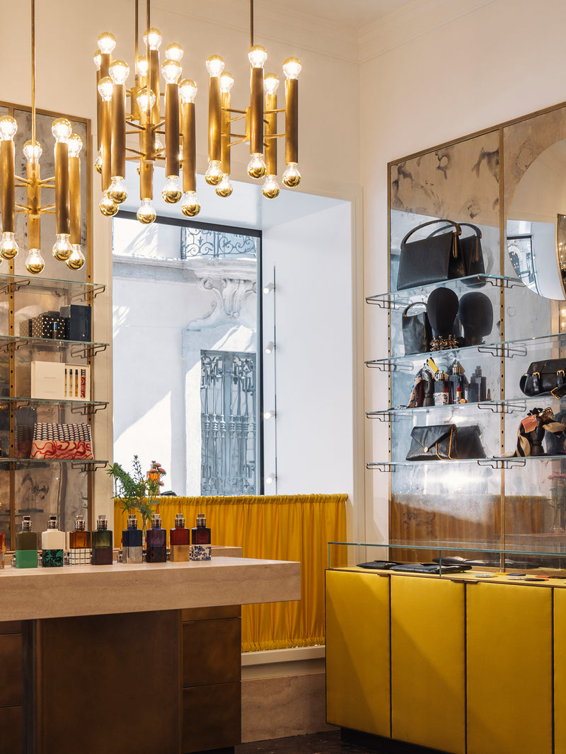 A luxury retail interior featuring a prominent gold multi-arm chandelier, yellow display cabinets, marble shelving, and handbag racks.