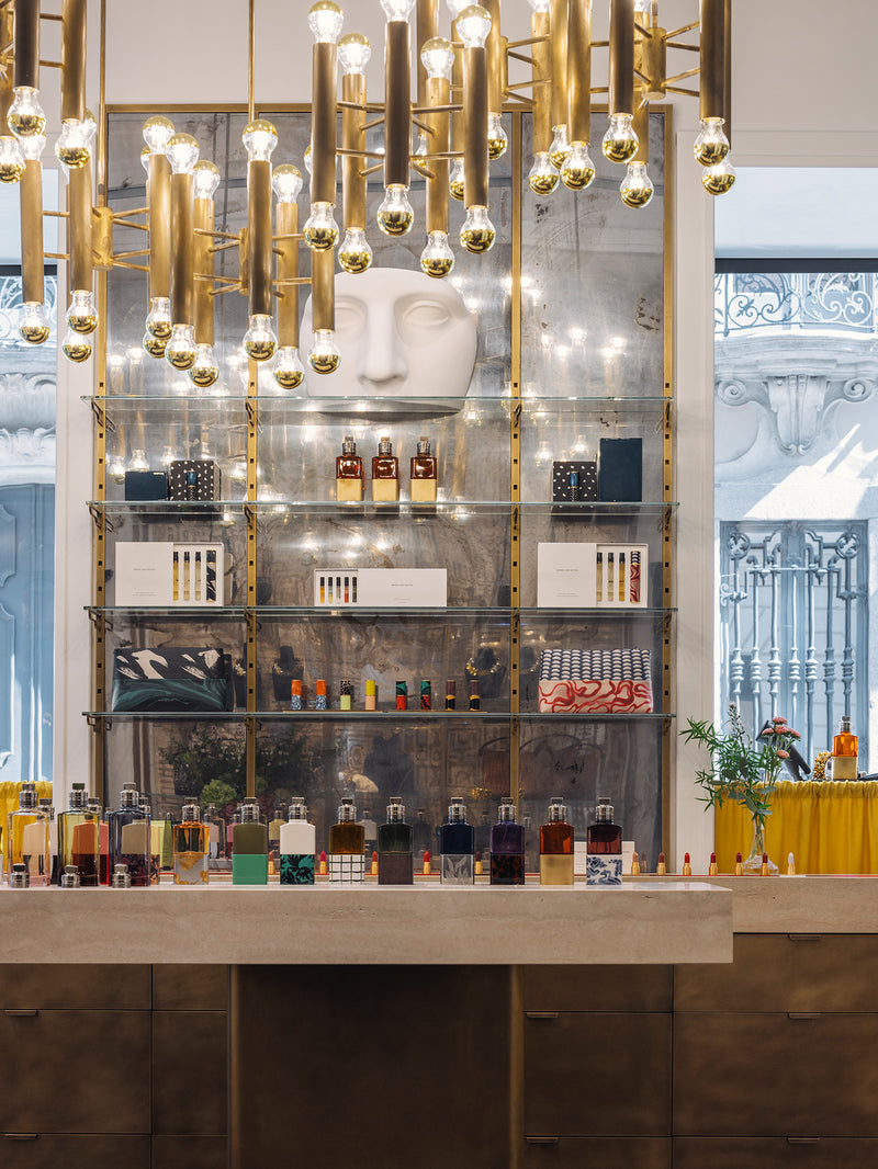 A luxury display featuring a large white sculptural face mounted on a glass shelving unit with gold-framed metal bars. Numerous fragrance bottles and make-up accessories are neatly arranged on the shelves and the marble countertop below.
