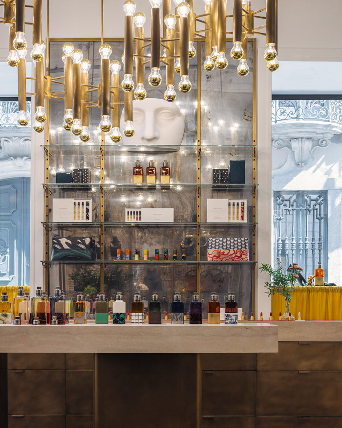 A luxury display featuring a large white sculptural face mounted on a glass shelving unit with gold-framed metal bars. Numerous fragrance bottles and make-up accessories are neatly arranged on the shelves and the marble countertop below.