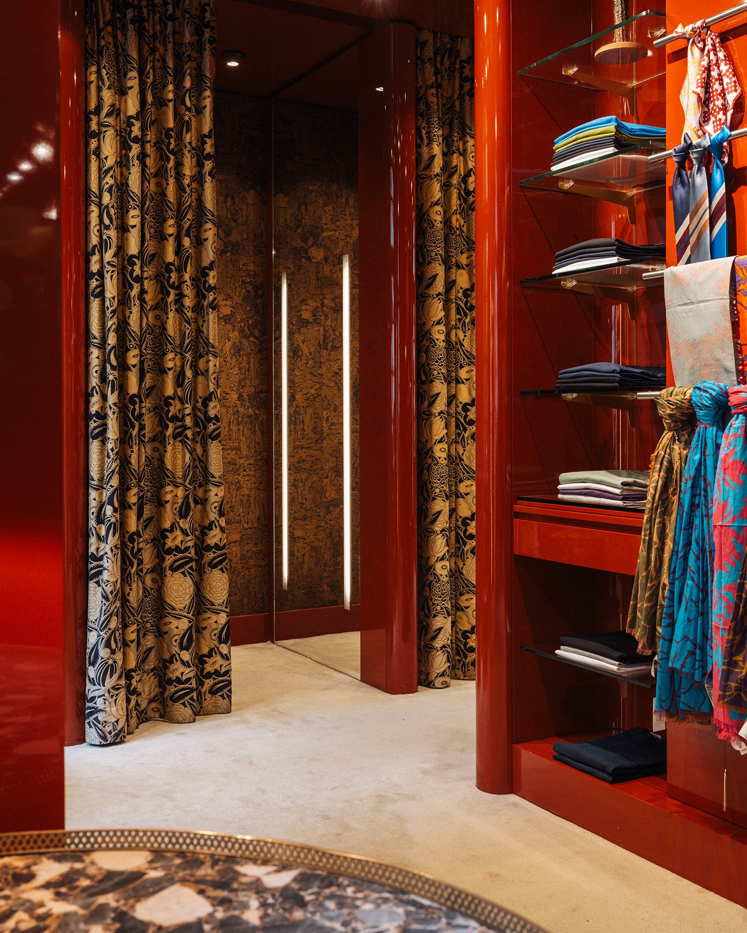 A luxury fitting room with glossy red wood walls, patterned beige and black curtains, and built-in shelving holding folded clothing and scarves.