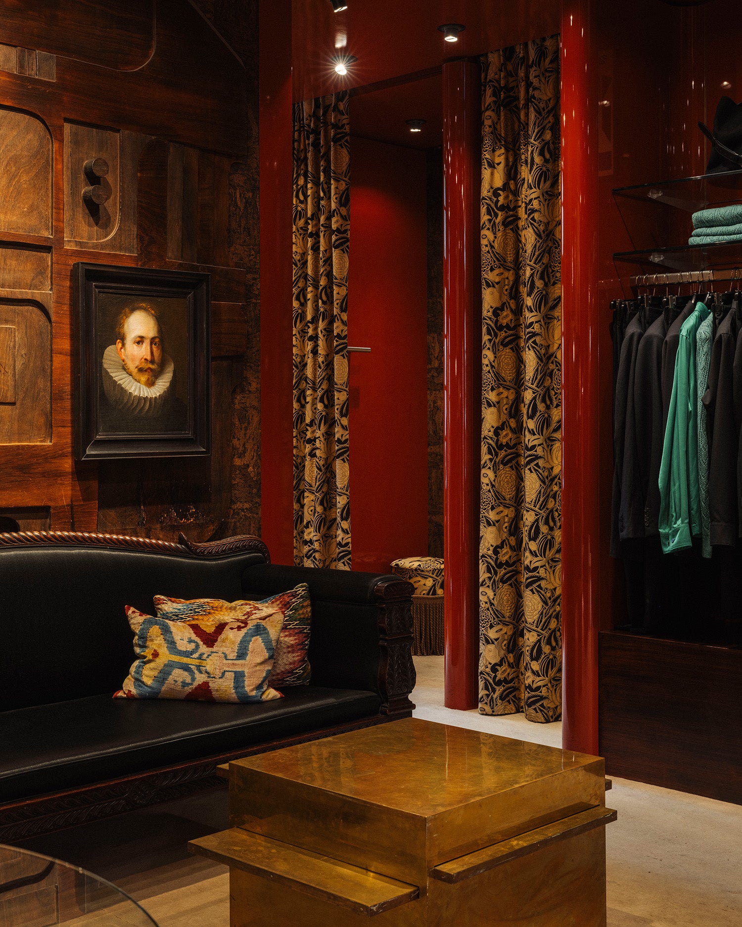 An elegant interior space with dark wood paneling, a black leather sofa with a patterned pillow, a wooden coffee table, and a clothing rack with green and black garments. A framed portrait hangs on the left wall.
