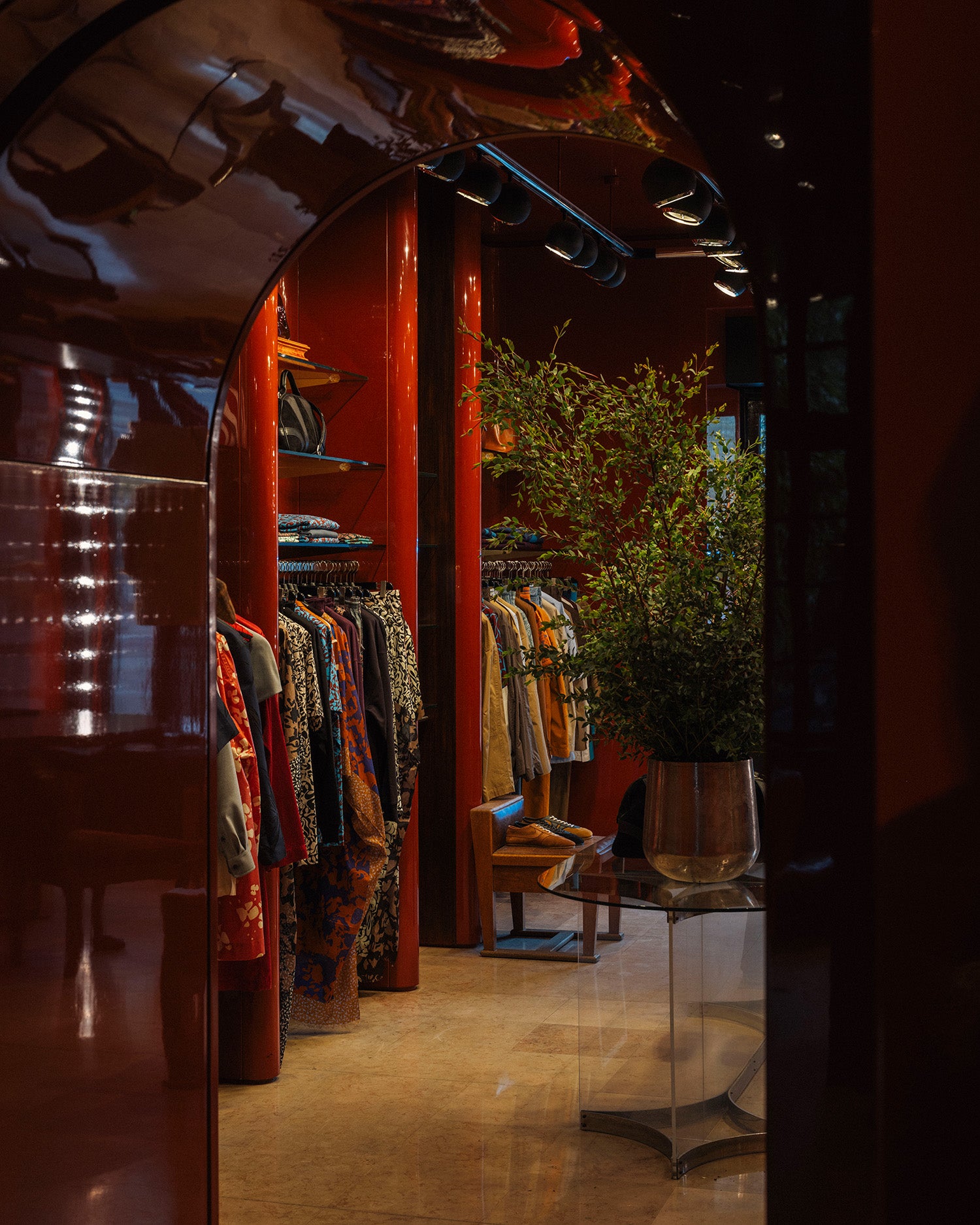 A boutique interior with warm red walls, clothing racks filled with garments, a green plant, and a reflective dark archway framing the scene.