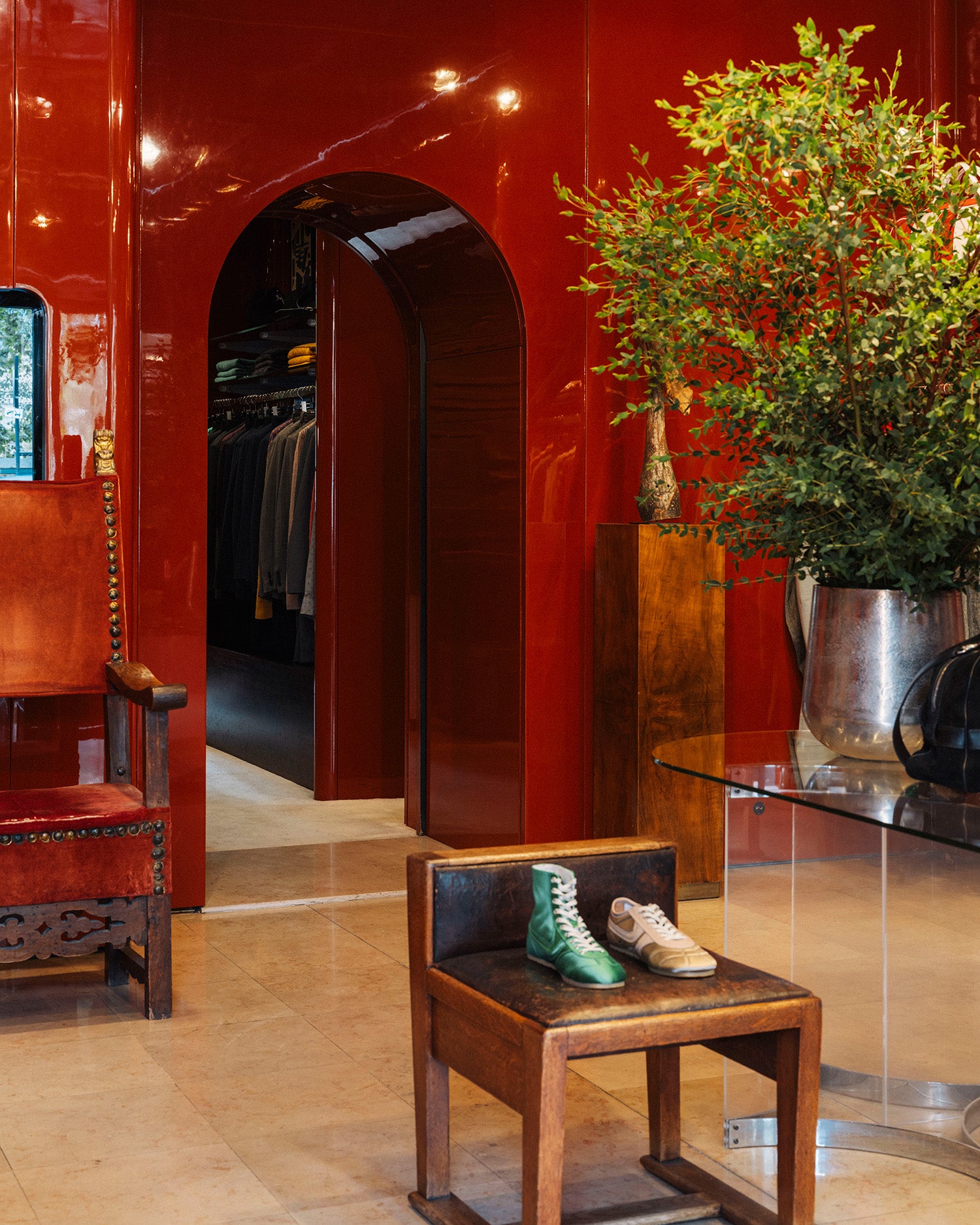 A styled interior scene with glossy deep red walls, an open arched doorway revealing clothing racks, a small wooden stool with a green and beige shoe on top, and a large plant on a glass table.